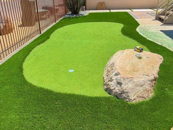 An artificial turf putting green featuring a large rock and surrounded by a fence.