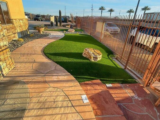 A walkway with artificial turf and rock accents, bordered by a metal fence and illuminated by the setting sun.